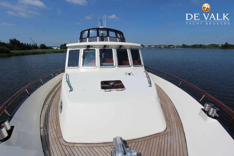 Een witte motorboot met een houten dek vaart op een waterweg met rietkragen en bebouwing op de achtergrond.