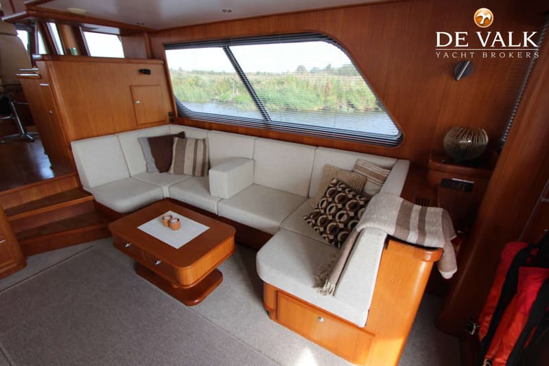 Een interieur van een boot met een L-vormige bank, een salontafel en een raam met uitzicht op water en riet.