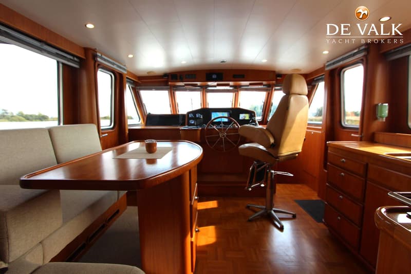 De foto toont het interieur van een boot met een houten tafel, banken, een stuurstand en een stuurstoel.
