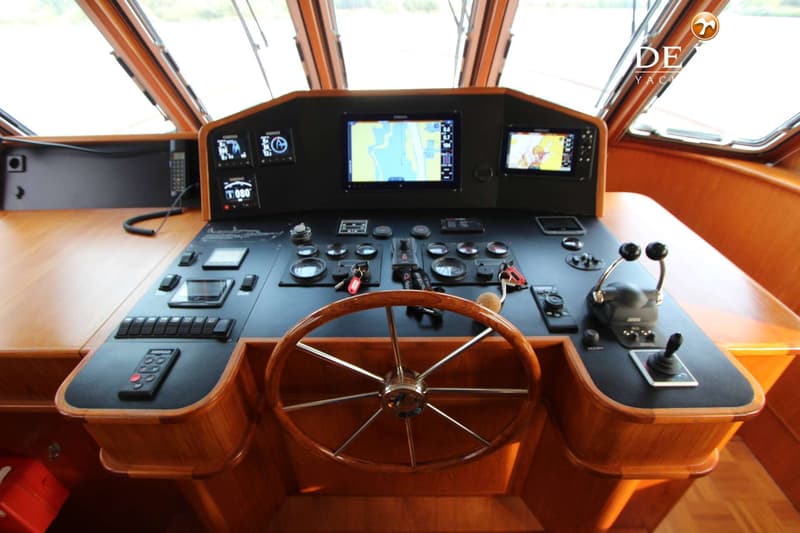Een houten stuurwiel en een zwart dashboard met diverse schermen, knoppen en hendels zijn zichtbaar in de stuurhut.