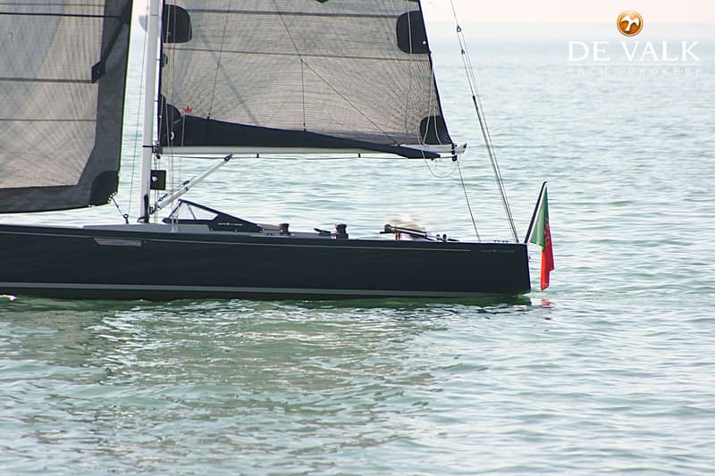 Een donkergrijze zeilboot met zwarte zeilen en een Portugese vlag aan de achtersteven vaart op kalm water.