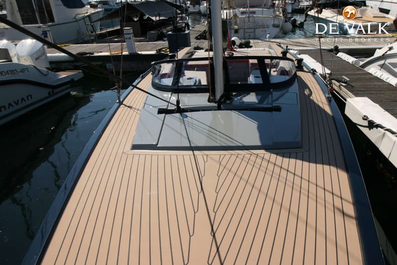 Een boot met een lichtbruin dek met zwarte strepen, een grijze opbouw en een mast, liggend aan een steiger.