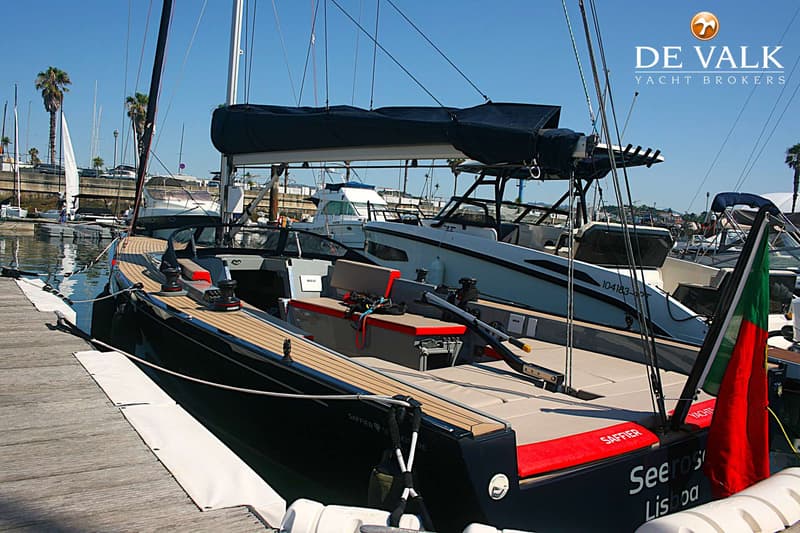 Een zwarte zeilboot met teakhouten dek ligt afgemeerd aan een houten steiger, met een Portugese vlag aan de achtersteven.