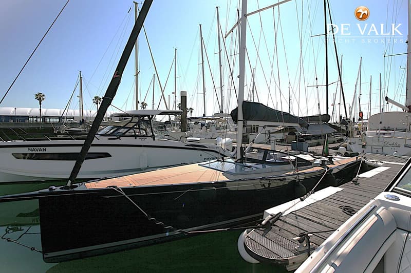 Een donkere zeilboot met houten dek ligt afgemeerd aan een steiger, met andere boten op de achtergrond.