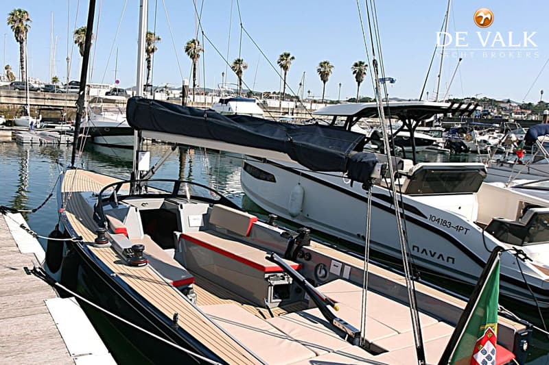 Een zeilboot met een houten dek en beige kussens ligt afgemeerd aan een steiger, met een andere boot ernaast.