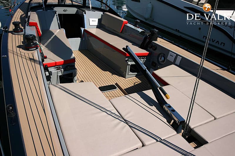 De foto toont het teakdek van een boot met grijze en rode kussens, twee zwarte Harken lieren en een zilverkleurige giek.