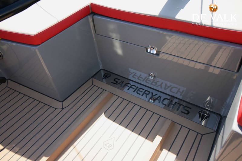 Een detailfoto van een bootinterieur met een grijze wand, rode en witte kussens, en een houten vloer met het logo…