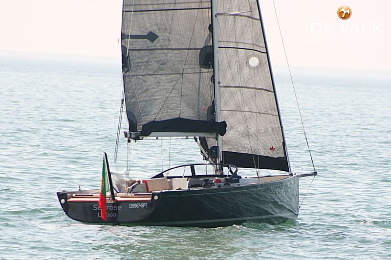 Een donkergrijze zeilboot met zwarte zeilen vaart op kalm water, met een Portugese vlag aan de achtersteven.