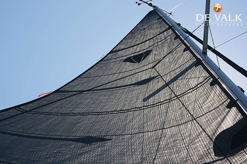 Een donker zeil met een ruitpatroon en een mast, gezien vanuit een laag perspectief tegen een helderblauwe lucht.