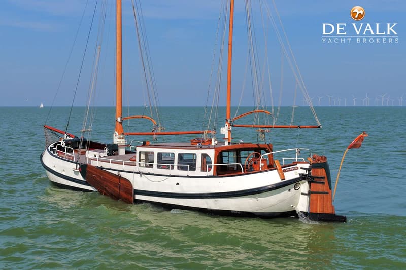 Een witte boot met houten accenten en twee masten vaart op groen water, met windmolens op de horizon.