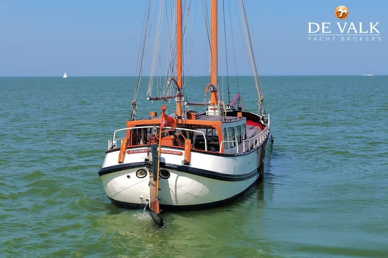 Een witte boot met oranje masten en de naam "STAVOREN" op de boeg vaart op rustig groen water. — Tjalk Koopmans Welvaren