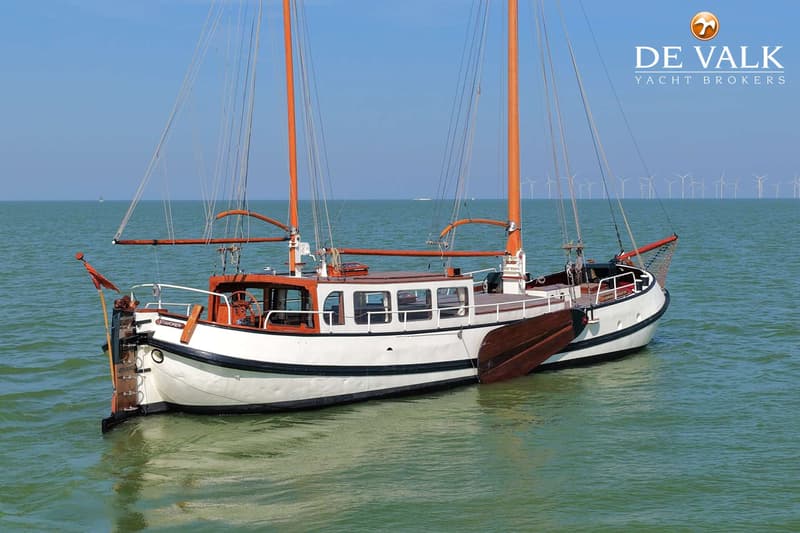 Een witte zeilboot met oranje masten en bruine accenten ligt op het water, met windmolens op de achtergrond.