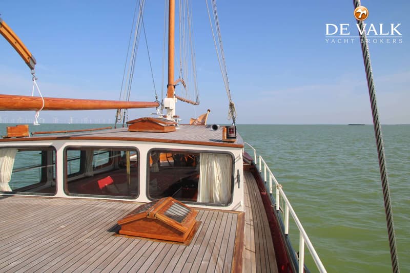 Een houten zeilboot met een kajuit en een mast, varend op groen water met windmolens aan de horizon.
