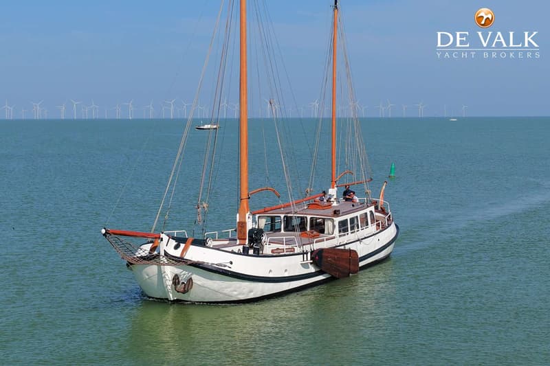 Een witte zeilboot met twee masten en bruine accenten vaart op rustig water, met windmolens op de horizon.