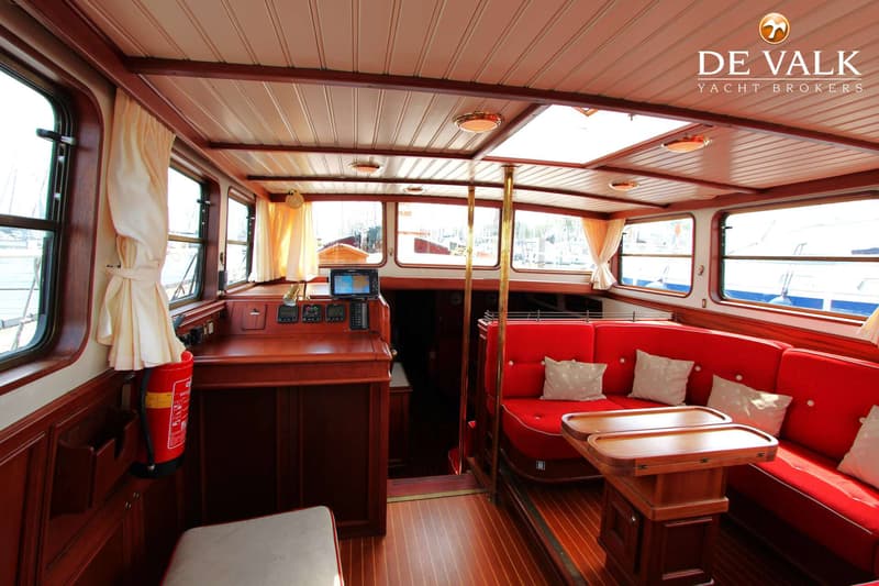 De foto toont het interieur van een boot met houten lambrisering, rode banken, een tafel en ramen met crèmekleurige…