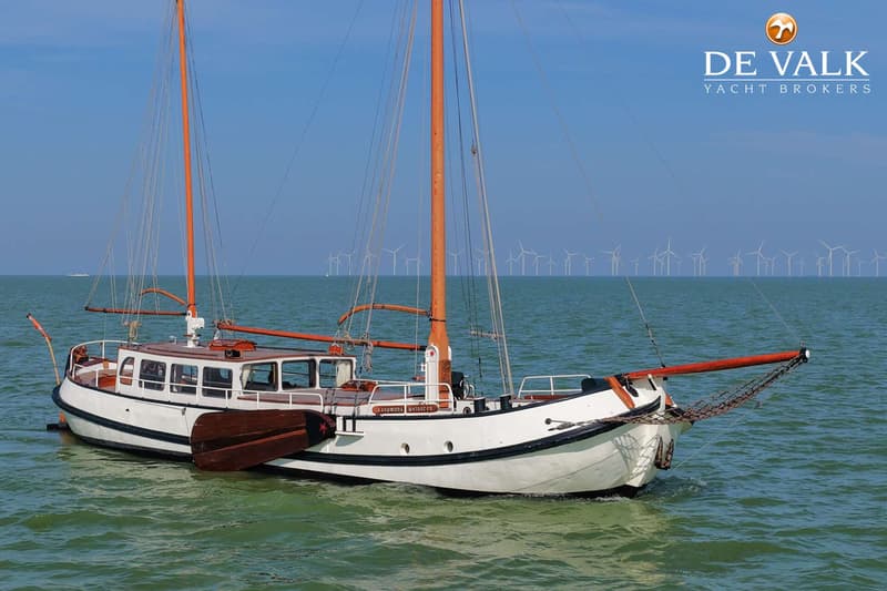 Een witte zeilboot met twee oranje masten vaart op groen water, met een windmolenpark op de achtergrond.