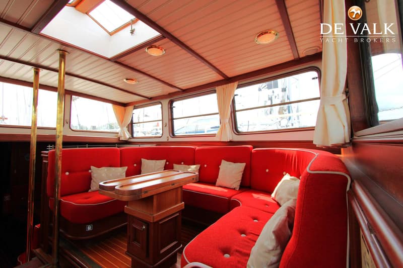 De foto toont het interieur van een boot met rode banken, een houten tafel, ramen met gordijnen en een dakraam.