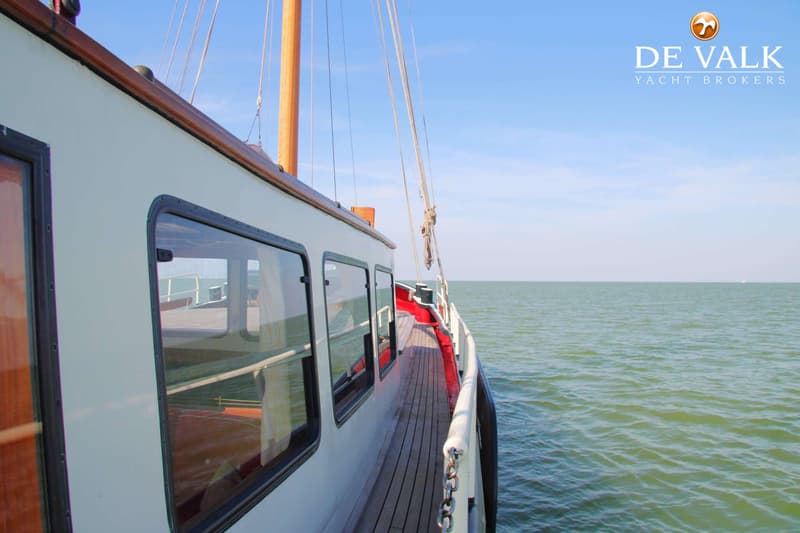 De foto toont de zijkant van een boot met ramen en een houten dek, varend op open water onder een blauwe lucht.
