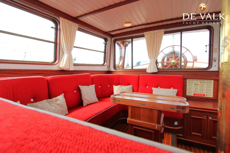 Een interieur van een boot met rode banken, een houten tafel en ramen met crèmekleurige gordijnen. — Tjalk Koopmans Welvaren