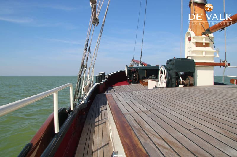 Een houten dek van een schip met een mast, lieren en reling, varend op open water onder een blauwe lucht.