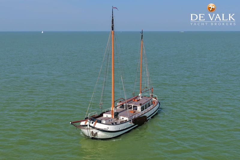 Een witte tweemast zeilboot met houten dek en oranje masten vaart op groen water onder een heldere hemel.