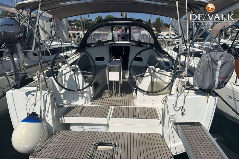 De foto toont de achterkant van een zeilboot met twee stuurwielen, een houten dek en een grijze buiskap.