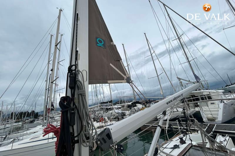 Een zeilboot met een opgerold zeil en diverse lijnen, liggend in een haven vol met andere zeilboten onder een bewolkte hemel.