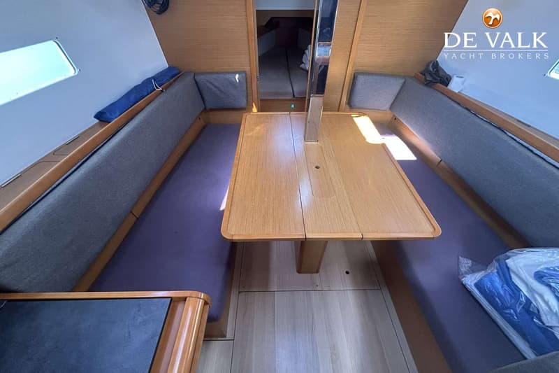 De foto toont het interieur van een boot met een houten tafel, banken met grijze kussens en een doorgang naar een…