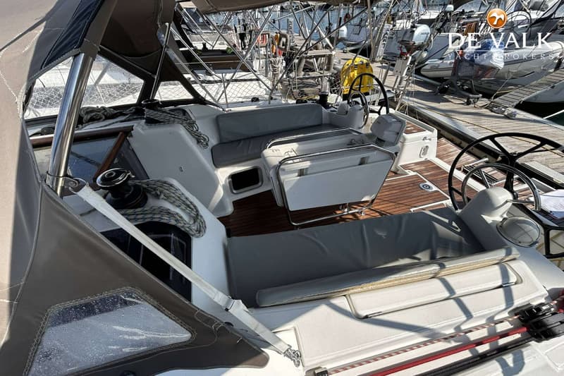 De foto toont de kuip van een zeilboot met teakhouten vloer, grijze kussens, twee stuurwielen en een opklapbare tafel.