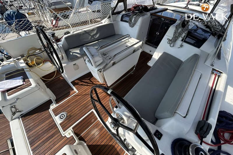 De foto toont de kuip van een zeilboot met een teakhouten dek, twee stuurwielen en zitbanken met grijze kussens.
