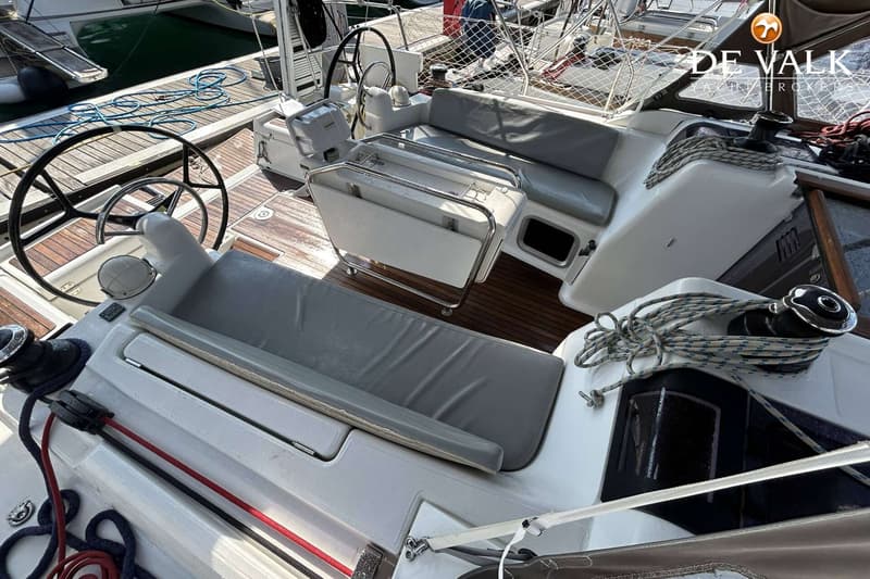 Een bovenaanzicht van de kuip van een zeilboot met twee stuurwielen, zitbanken met grijze kussens en een opklapbare tafel.