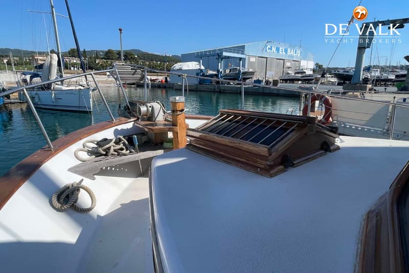 De foto toont het voordek van een boot met een houten luik, touwen en een ankerlier, met op de achtergrond een haven.