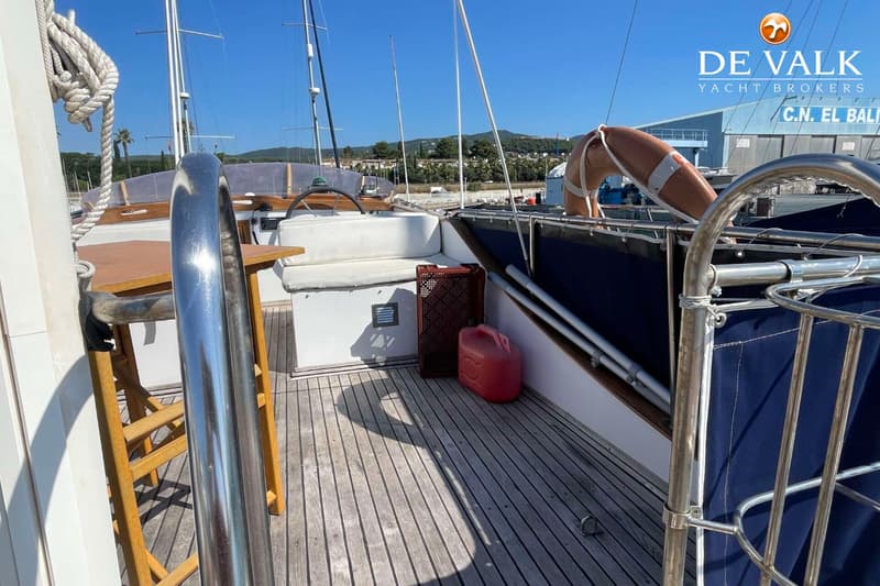 De foto toont het teakhouten dek van een zeilboot met een stuurwiel, witte kussens, een houten tafel en een rode jerrycan.