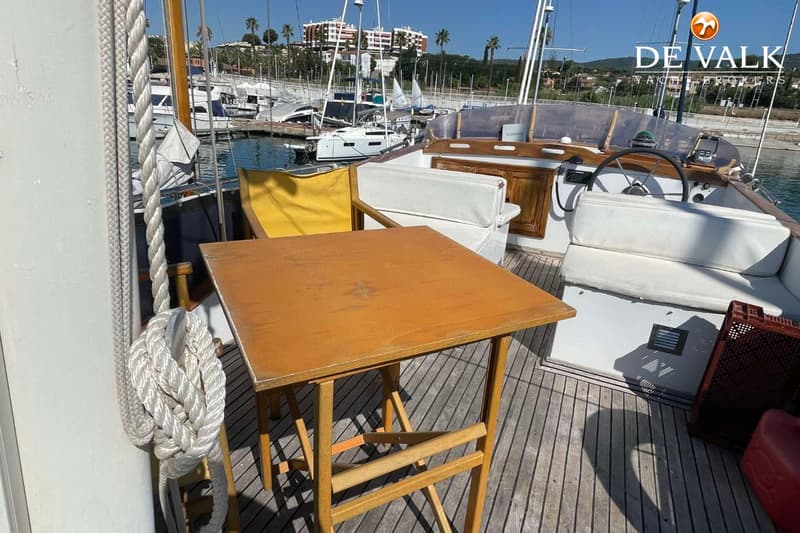 Een houten tafel en een gele stoel staan op het dek van een boot, met een haven op de achtergrond.