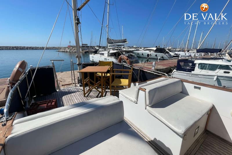 Een dek van een boot met witte kussens, een houten tafel en stoelen, en een reddingsboei, met een jachthaven op de…