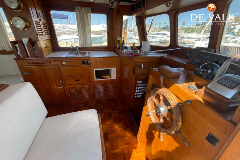 De foto toont het interieur van een boot met een houten stuurwiel, een kombuis met spoelbak en magnetron, en een bank met…