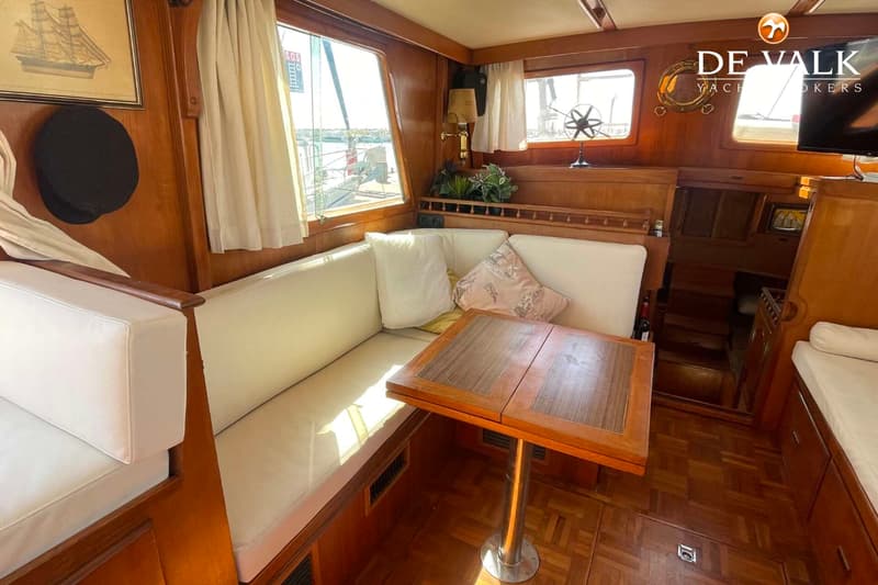 De foto toont een houten interieur van een boot met een bank, tafel, ramen en een schilderij.
