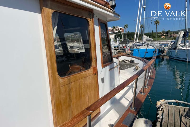 Een witte boot met houten details en ramen ligt in een haven, met andere boten en gebouwen op de achtergrond.