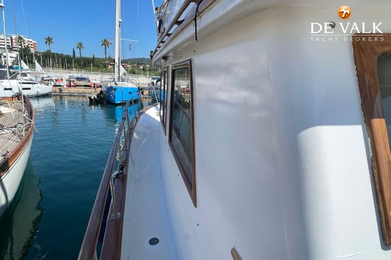 De foto toont de zijkant van een witte boot met houten details, liggend in een haven met andere boten en palmbomen op de…