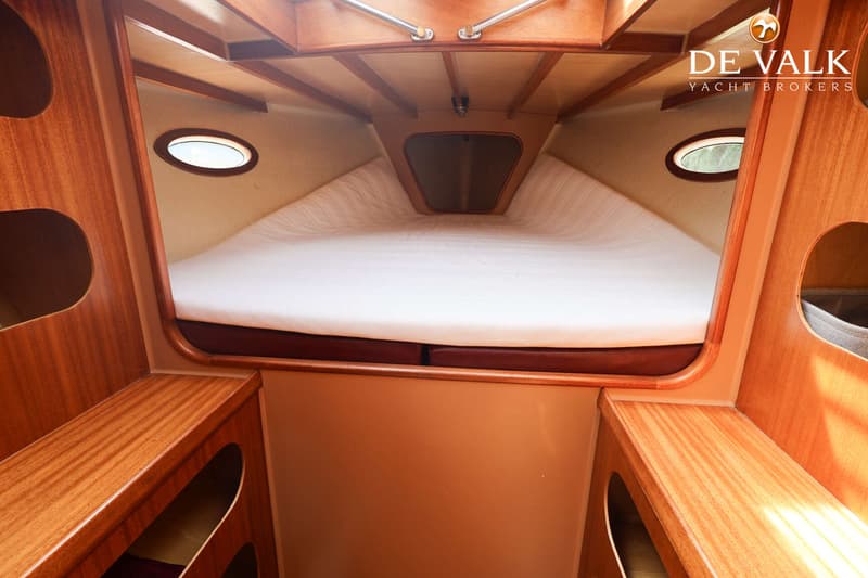 De foto toont het interieur van een kajuit met een V-vormig bed, twee patrijspoorten en houten afwerking.