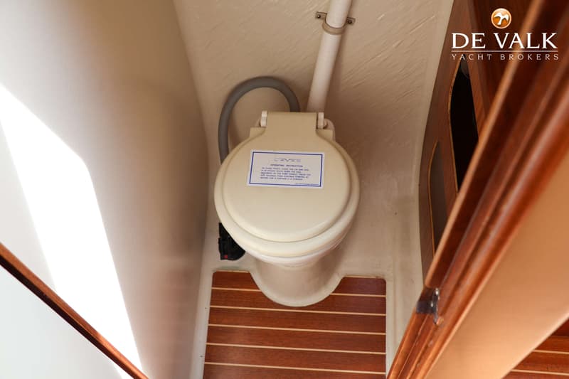 Een crème toilet met een blauw-wit instructielabel op de deksel staat op een houten vloer met lichte strepen.