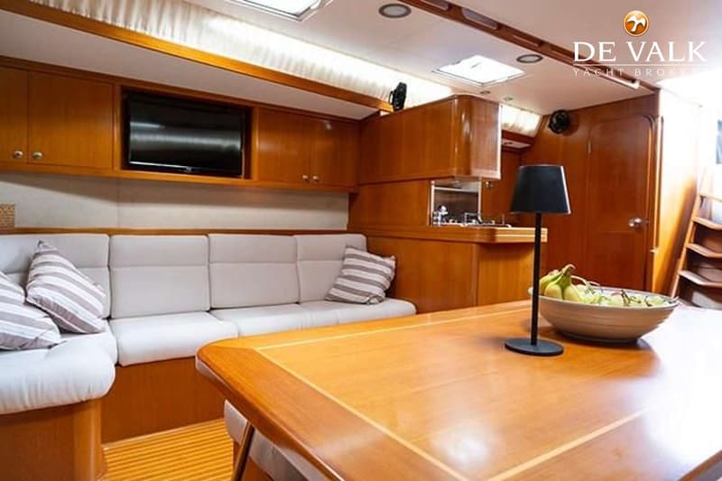 Een interieur van een boot met een houten tafel, banken met lichte kussens, een televisie en een kombuis. — Baltic 60