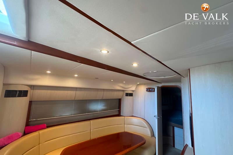 Een interieur van een boot met een U-vormige beige bank, een houten tafel en een open deur naar een andere ruimte.