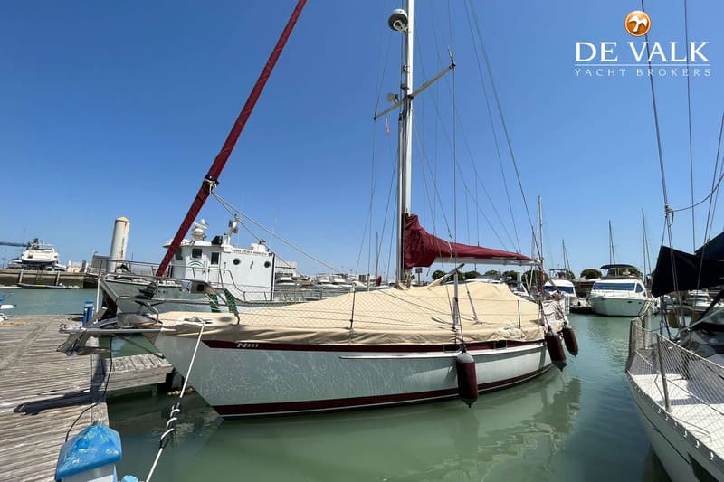 Een witte zeilboot met rode strepen en een beige afdekzeil ligt aangemeerd in een haven onder een helderblauwe lucht.