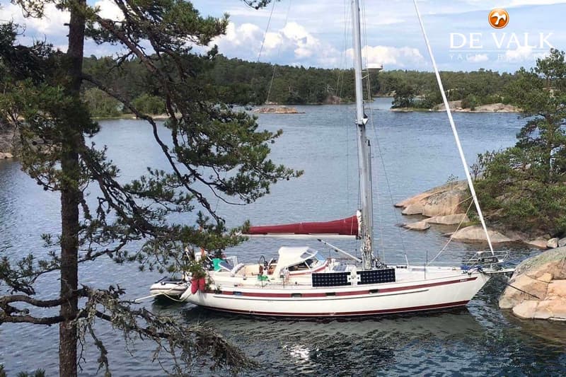 Een witte zeilboot met rode strepen en zonnepanelen ligt in een baai met rotsachtige oevers en bomen. — Najad 391