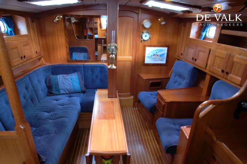 De foto toont een houten interieur van een boot met blauwe gestoffeerde banken en stoelen, een tafel en diverse decoraties.