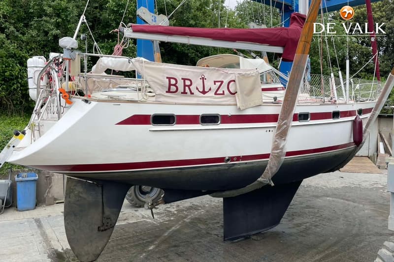 Een witte zeilboot met rode strepen en de naam "BRIZO" op een doek, hangt in een takel boven een betonnen ondergrond.