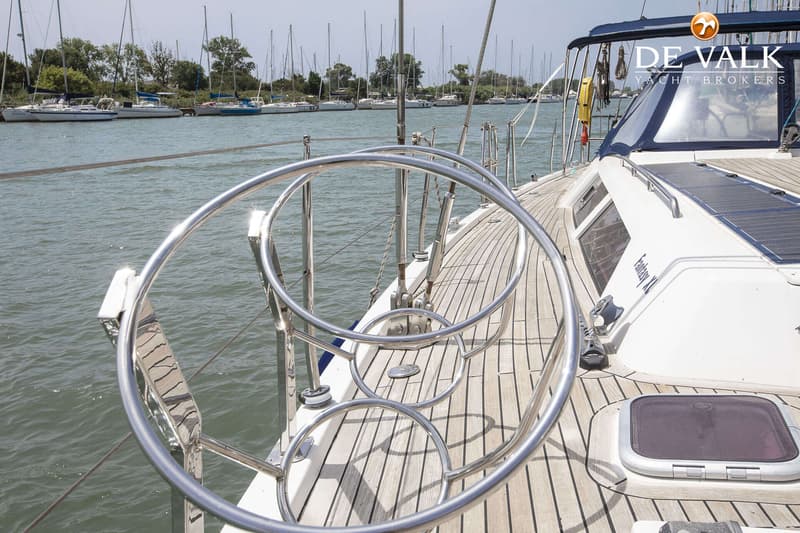 Een zeilboot met een teakhouten dek en een rvs-reling, liggend aan een kade met andere boten op de achtergrond. — Hanse 540e
