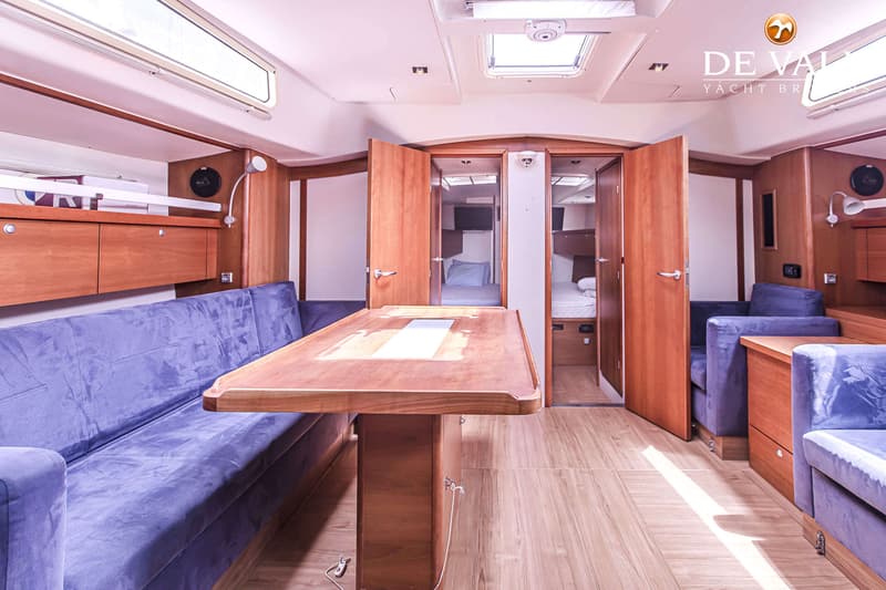 De foto toont het interieur van een boot met een houten tafel, twee blauwe banken en twee open deuren naar slaapcabines.