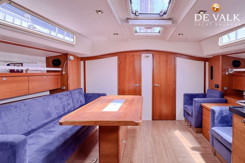 De foto toont het interieur van een boot met een houten tafel, blauwe banken, houten kasten en deuren, en ramen in het…